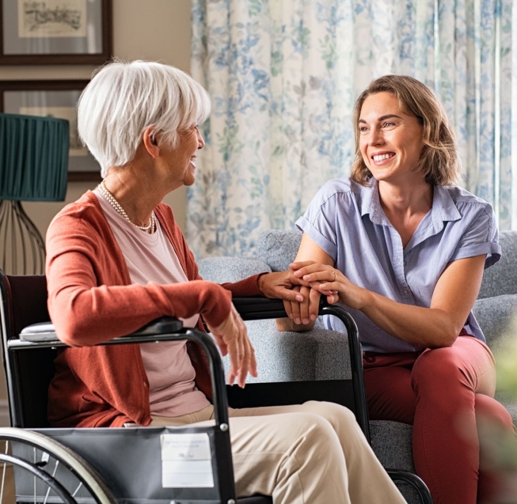 Caregiver sitting beside an elderly woman in a wheelchair while holding her hand and smiling during a conversation.