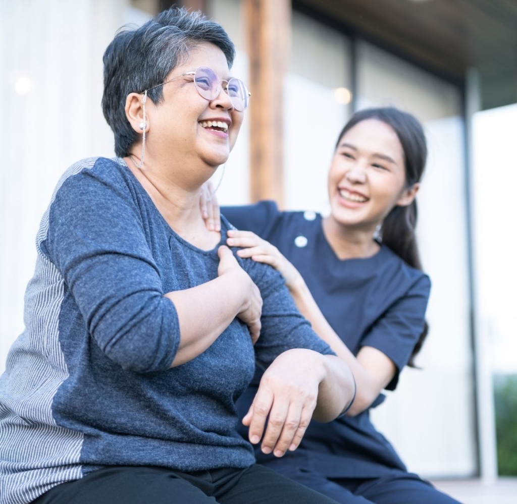Caregiver smiling and touching the shoulder of an elderly woman as they sit outside and share a joyful moment.