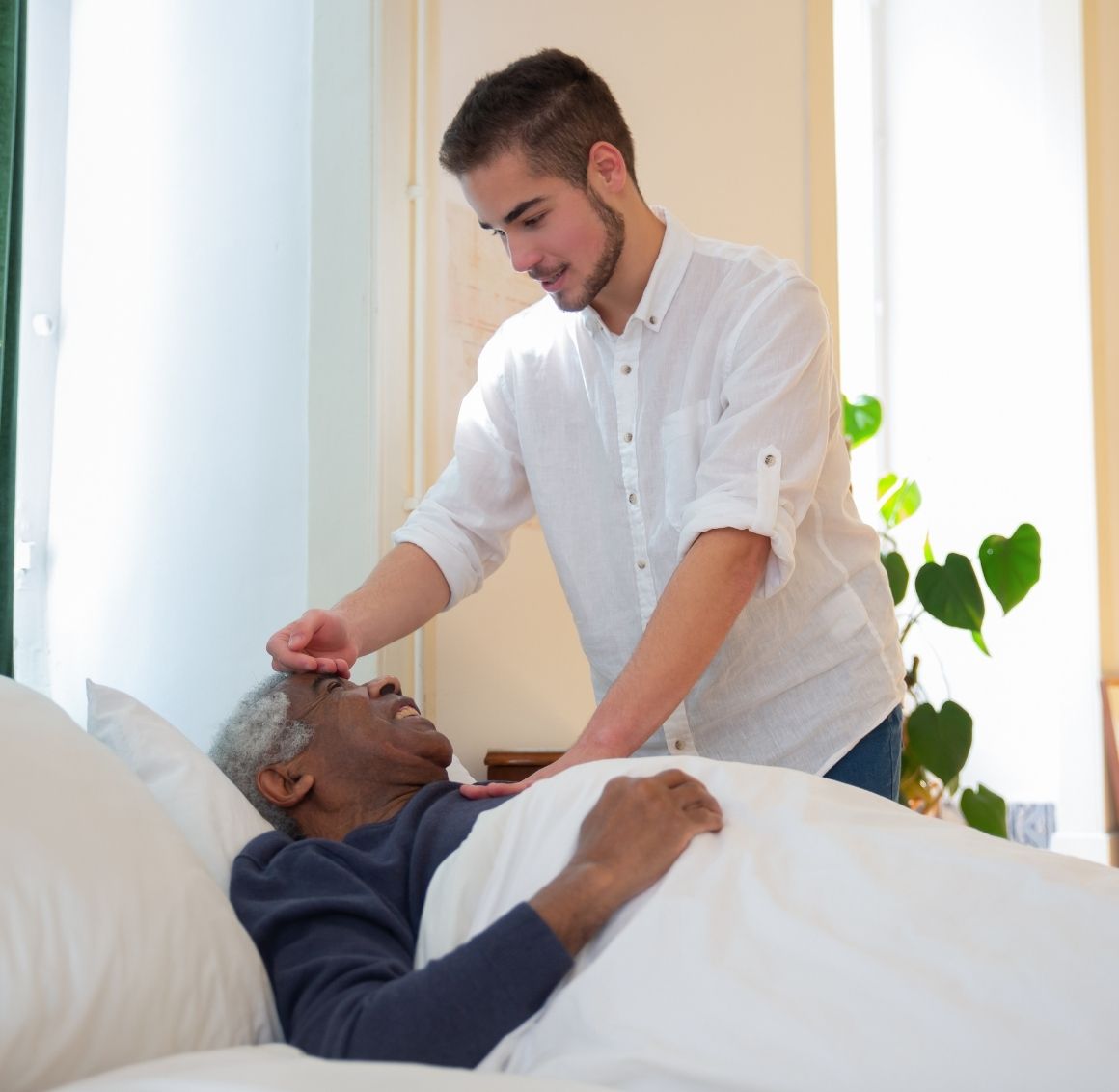 Caregiver helping an elderly man rest comfortably in bed, gently checking on him in a bright and calm home setting, representing 24 Hour & Live In Care.
