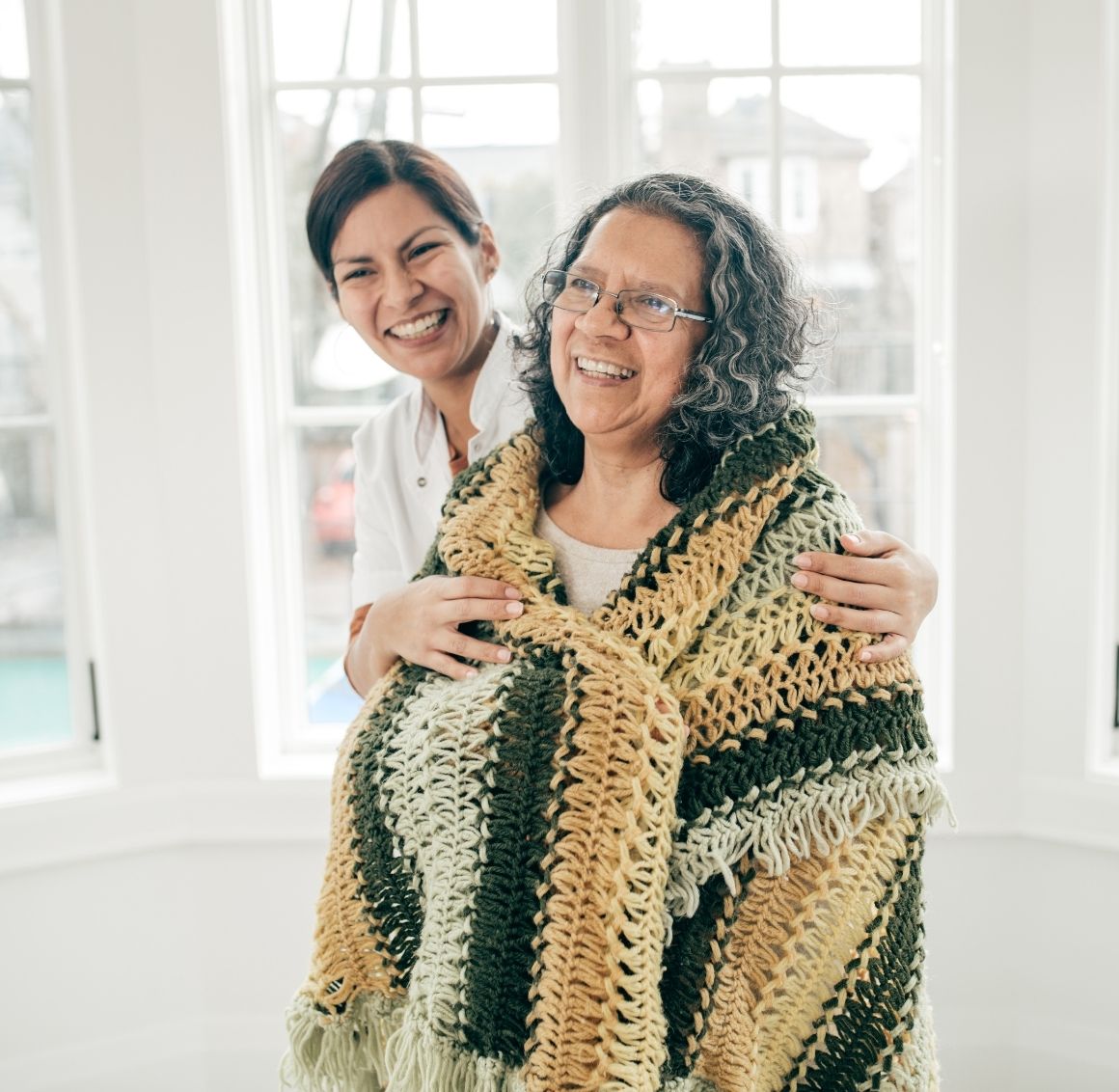 Caregiver wrapping a warm knitted blanket around a smiling elderly woman near bright windows symbolizing comfort and emotional support.