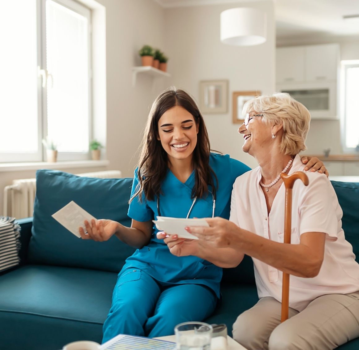 Caregiver and elderly woman sitting on a couch smiling and reviewing papers together in a warm and inviting living room.