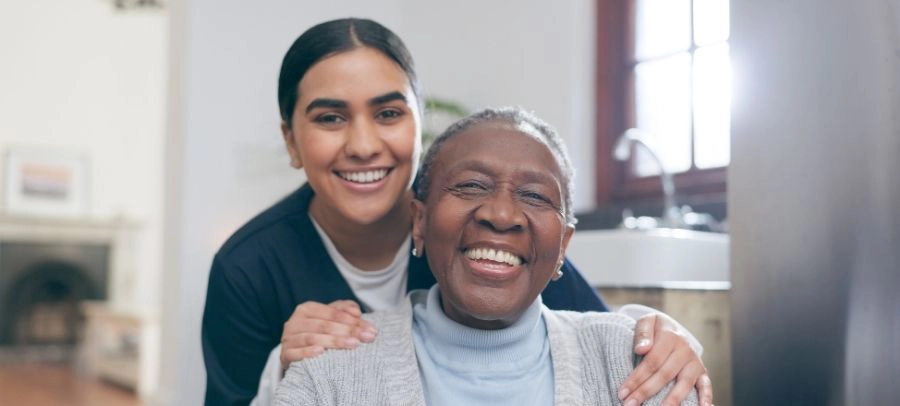 Smiling caregiver hugging an elderly woman from behind in a bright home environment showing warmth and emotional support.