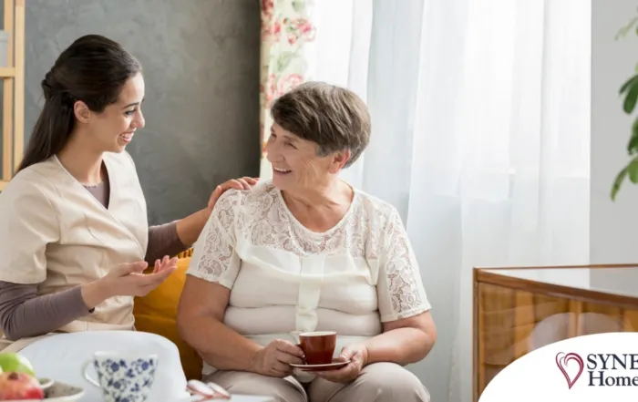 Smiling caregiver with her hand on an older woman’s shoulder as they share tea on the couch, showing home care vs assisted living