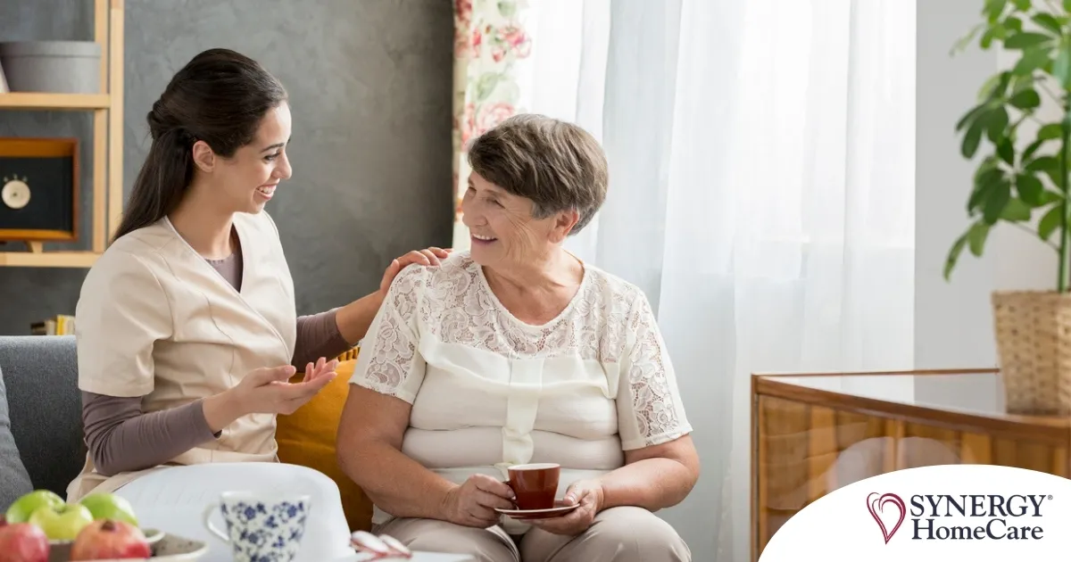 Smiling caregiver with her hand on an older woman’s shoulder as they share tea on the couch, showing home care vs assisted living